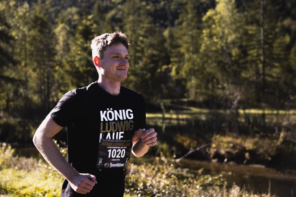Ein lächelnder Läufer im schwarzen König-Ludwig-Lauf-Shirt läuft im Sonnenlicht.