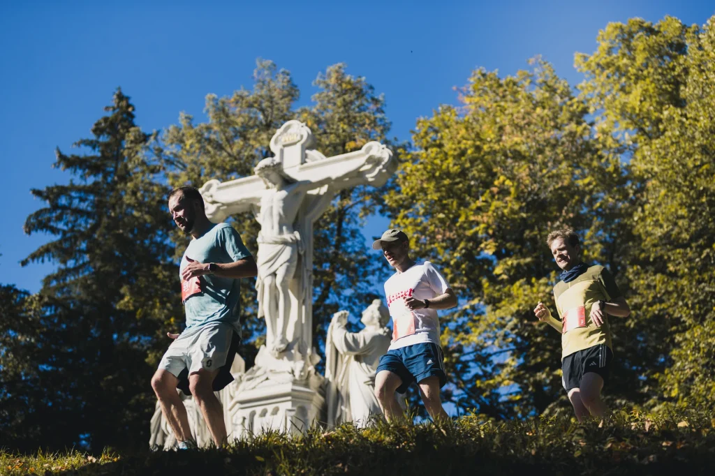 Laufende passieren eine steinerne Kreuzigungsgruppe unter blauem Himmel.