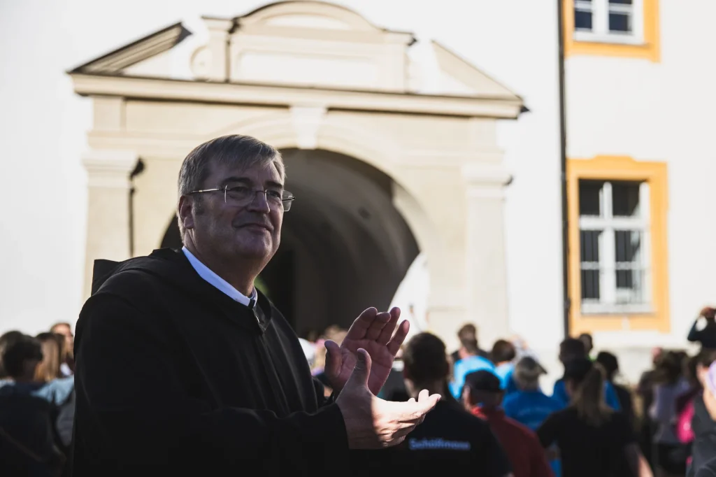 Ein lächelnder Mann klatscht vor einem Gebäude des Klosters Ettal; weitere Menschen stehen im Hintergrund.