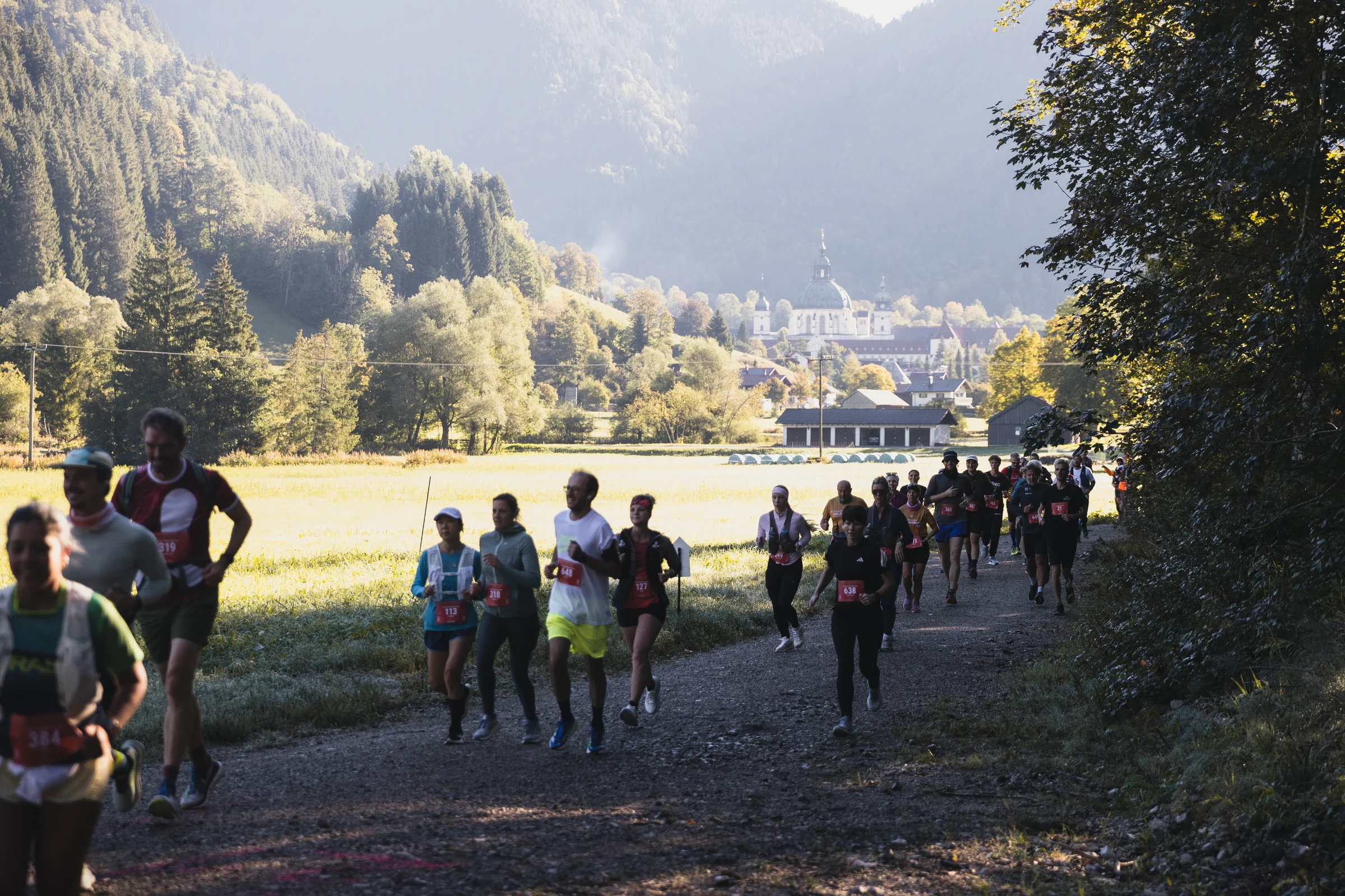 Eine Laufgruppe auf einem Weg mit sonnigen Wiesen und dem Kloster Ettal im Hintergrund.